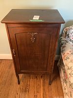 Front view of vintage mahogany record cabinet showing wood finish and ornate metal embellishment on door, with key lock and curved legs.