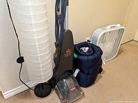 White cylindrical floor lamp, Bissell carpet cleaner, rolled blue sleeping bag, white box fan shown grouped on carpet floor against wall.