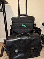 Front view of black makeup roll-around bag with extended telescoping handle and black leather briefcase satchel placed in front on the floor.