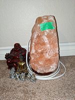 Photo showing a large Himalayan salt lamp with wooden base and six Buddha figurines of varying sizes and finishes arranged on carpeted floor near a wall.