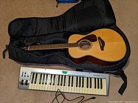 Overview of Yamaha acoustic guitar in black softshell case alongside M-Audio keyboard on carpet