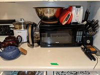 Countertop with Aroma rice cooker and glass lid, Farberware kettle, RCA microwave, two ceramic teapots (brown and blue), stainless steel mixing bowl, red silicone oven mitts, and knife block with knives.