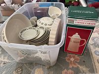 Plastic bin filled with various Christmas-themed china dishes including plates, bowls, and serving pieces. Also shows the boxed coffee carafe next to the bin.