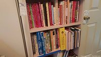 Shelf with about 60 cookbooks, both hardcover and softcover, colorful and plain covers, arranged standing upright.