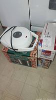 White Oster Belgian waffle maker with black cord placed on boxes with visible branding on floor.