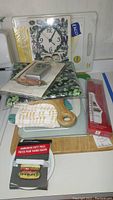 Photo showing six cutting boards stacked, celery spoon rest in packaging, new striped dish towels, chocolate paddle thermometer, hamburger press, baked potato bag, and small casserole dishes on a kitchen counter.
