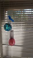 Two blown glass balls hanging in front of window blinds, one blue with green and white swirls, the other red with white streaks, suspended by string with a blue plastic piece.