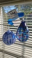 Two decorative glass balls hanging against blinds, one teardrop-shaped and multicolored, the other round with blue and white spiral swirl design.