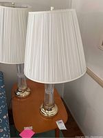 Two matching lamps on a wooden table, showing brass-like bases, clear ribbed glass stems, and white pleated lampshades.