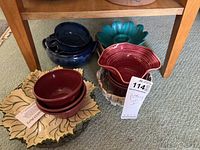 Photo of various pottery pieces including a dark blue bowl with handle and lid, turquoise flower-shaped plate, maroon bowls on a decorative leaf-shaped plate, and a ceramic basket.