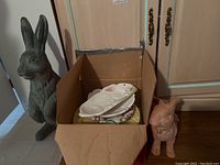 Large gray and smaller pink rabbit statues with opened cardboard box holding plates and bowls with rabbit or egg decorations