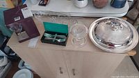 Wide view of lot items on cabinet including boxed wine glasses, green box with forks, clear glass serving dishes, large covered serving dish with wooden base