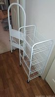 White 2-step ladder positioned next to white metal wire 3-tier shelf cart on a wooden floor.