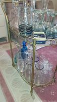 Side view of brass tea cart with glass shelves showing assorted glassware including glasses and decanters.