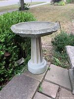 Side angle image showing full birdbath, fluted pedestal, and surrounding outdoor setting with some greenery and paving stones.