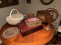 Full view of lot items arranged on round wooden table including white wicker basket, copper pot, decorative fan, glass platters, and boxes.