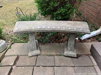 Concrete garden bench showing full front view of the curved top seat and two supporting legs placed outdoors on stone pavers near garden plants and red brick wall.