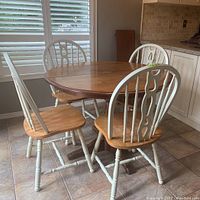 Round wooden kitchen table with four matching chairs arranged around it. Window shutters and kitchen cabinets in background.