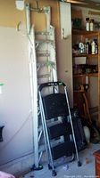 6-foot aluminum extension ladder leaning against a garage wall next to a smaller three-step foldable step ladder with black steps and handle, showing garage storage surroundings.