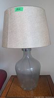Full view of a table lamp showing clear glass base and beige fabric shade