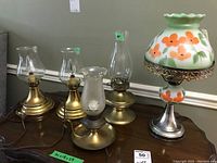 Five vintage lamps on a wooden table including brass and silver metal bases with clear and frosted glass chimneys and floral painted glass shade. All lamps vary in height from 9 to 17 inches.