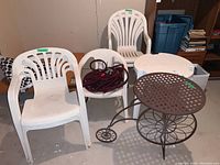 Front view of five white plastic patio chairs (three tall and two short), small round white plastic table, brown metal sculptural table, and garden hose coiled inside one chair.