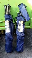 Two blue folding armchairs in carrying bags standing upright, showing the top metal frame parts and fabric bags.