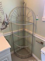 Front view of metal and glass corner shelving unit showing three glass shelves and metal frame with decorative finial.