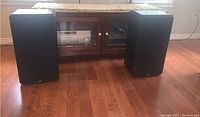 Two black Boston HD9 speakers placed side by side on a wooden floor, with a stereo receiver and glass-front console in the background.