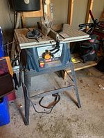 Full view of Ryobi 10 inch table saw on metal stand in storage with attached blade guard and miter gauge, showing overall condition and stand.