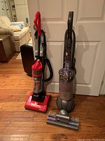Two upright vacuum cleaners standing side by side, Dirt Devil on left and Dyson on right, on wooden floor against white door.