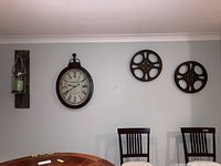 Four pieces of wall art hanging on a wall including two metal movie reels, a clock, and a barn board with glass jar and candle.