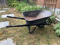Side angle image showing the full metal wheelbarrow with single wheel, black plastic grips on handles and surface rust inside the tub.