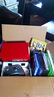 Box containing red Nintendo Wii console, black/blue controller, and several game/DVD cases.