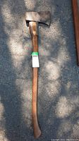 Full view of axe showing metal head with steel brace and full length wooden handle on asphalt surface with shadows.