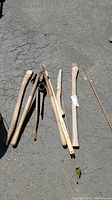 Multiple wooden axe handles, a pick axe head, thin metal pry bar, and a long branch trimmer pole laid on asphalt surface.