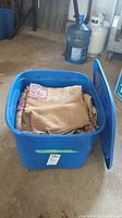 Blue plastic bin filled with folded burlap bags with visible printed text and partially attached lid