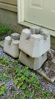Three concrete post supports stacked one on top of another placed near a door on a pebble and soil ground with some green plants.