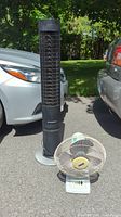 Front view of Honeywell HY-026 tower fan and Super Oscillating 12DFT desk fan placed outdoors between two cars.