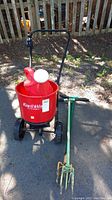 Overall view of the Earthway red walk-behind spreader with black handle and wheels, red hopper, and green hand cultivator next to it on pavement under sunlight.