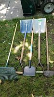 Full view of garden tools laid out on grass including rake, two shovels, ice chipper, and edger