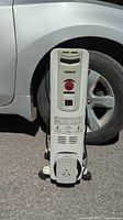 Full view of white Noma portable radiator standing upright with car tire background, showing control panel and base with cord wrap.
