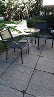 Two metal patio chairs with black mesh material seats and backs placed on paved patio area beside a metal round patio table with a glass top. Outdoor setting with greenery in background.