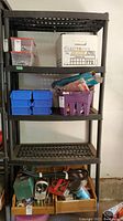 Full view of black plastic shelving unit containing various items. Shelving unit has four shelves and is sturdy.