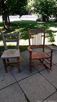 Two rustic wooden chairs outdoors on paving stones under shade of trees, one painted rocking chair and one natural wood chair.