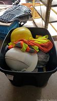 Bin full of hard hats including a white and yellow hard hat on top, along with fluorescent orange and yellow reflective vests and safety clothing inside a black plastic bin.