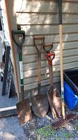 Photo showing four different shovels and a metal rake with wooden handle, leaning against a building.