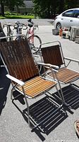 Two vintage aluminum frame wood slat folding lawn chairs set outdoors on pavement, one high back and one standard back