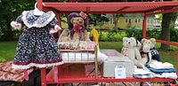 Four stuffed bears arranged with vintage style brass doll bed, floral dress hanging, and folded doll clothing on red perforated metal cart.