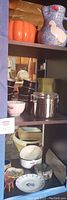 Black cubby showing upper shelves with large orange pumpkin pitcher and blue duck ceramic pitcher, metal cannister and other kitchen accessories like coasters and plates on lower shelves